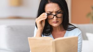 A middle-aged woman with glasses struggling to read her book.