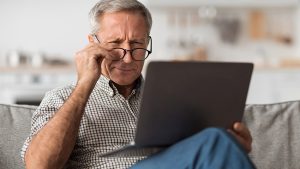 A senior man with glasses struggling to read on a laptop at home.